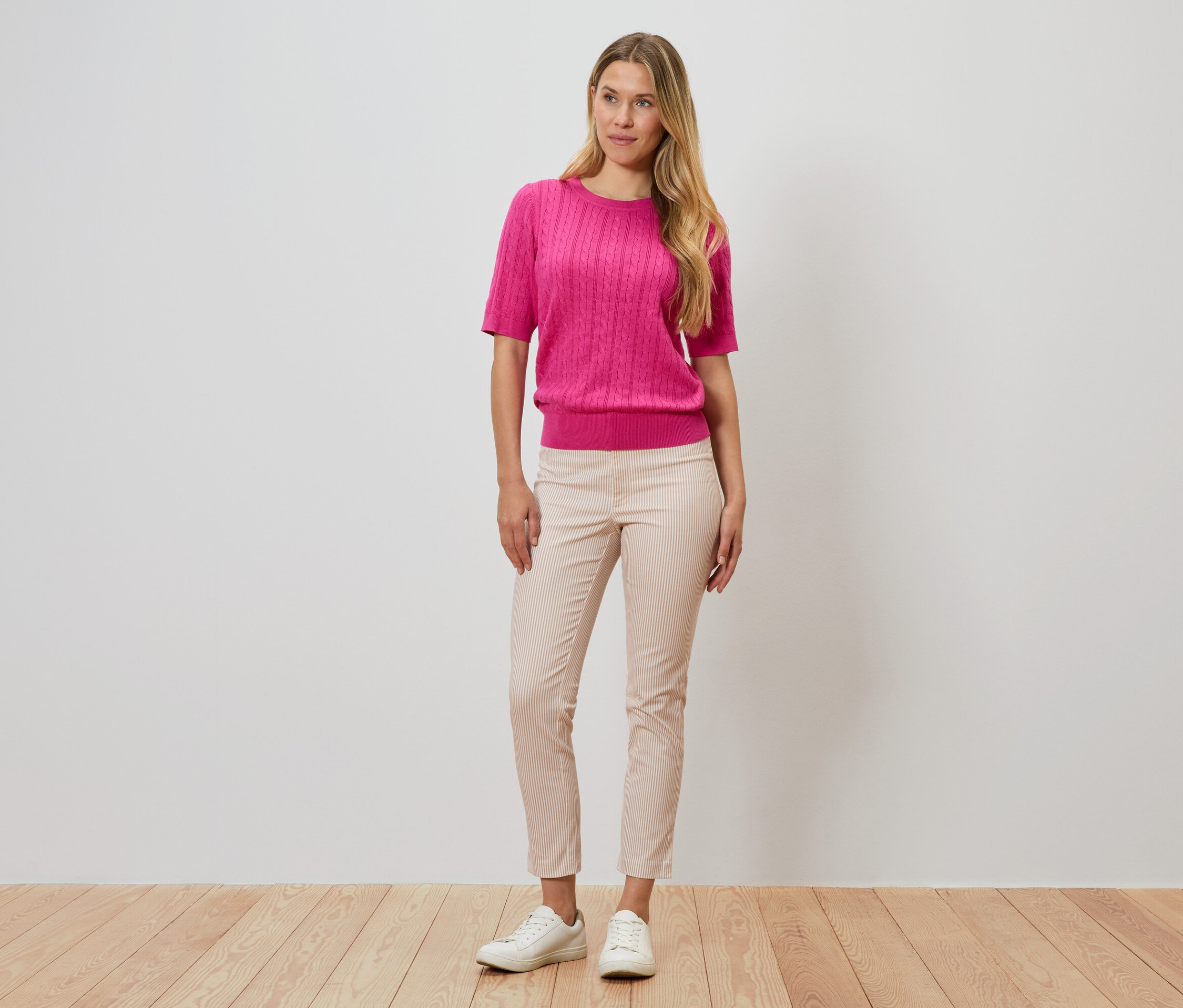 Kadın bir duvarın önünde duruyor, pembe bir kazak, çizgili pantolon ve beyaz ayakkabılar giyiyor.