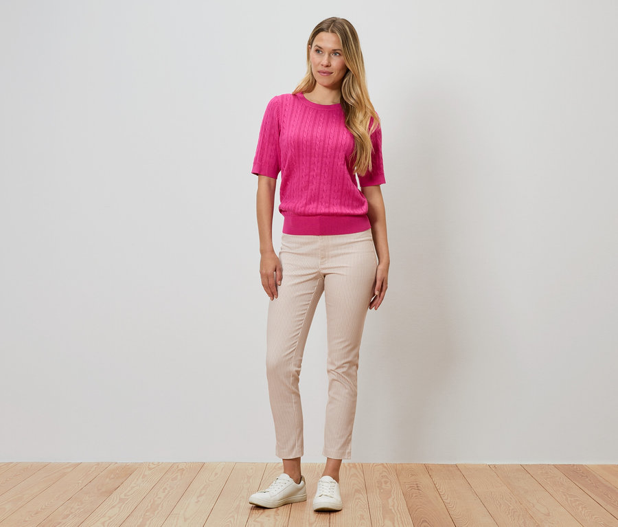 Kadın bir duvarın önünde duruyor, pembe bir kazak, çizgili pantolon ve beyaz ayakkabılar giyiyor.