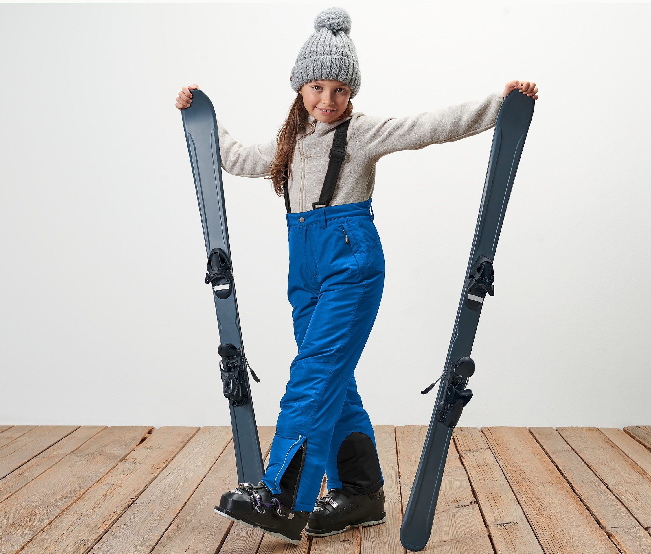 Gri şapkalı kız çocuğu kayakları tutuyor ve mavi çocuk kayak pantolonu giyiyor.