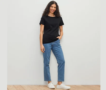 Siyah tişört, mavi kot pantolon ve beyaz ayakkabı giyen bir kadın beyaz bir duvarın önünde duruyor. 
