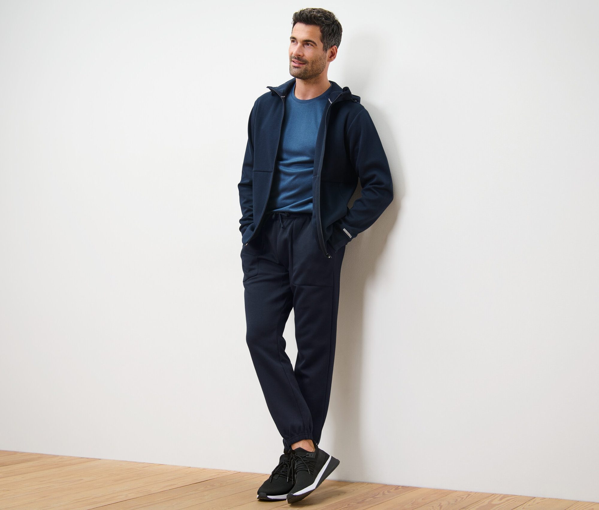 Duvara yaslanmış adam koyu lacivert kapüşonlu ceket, tişört ve fonksiyonel eşofman altı giyiyor.