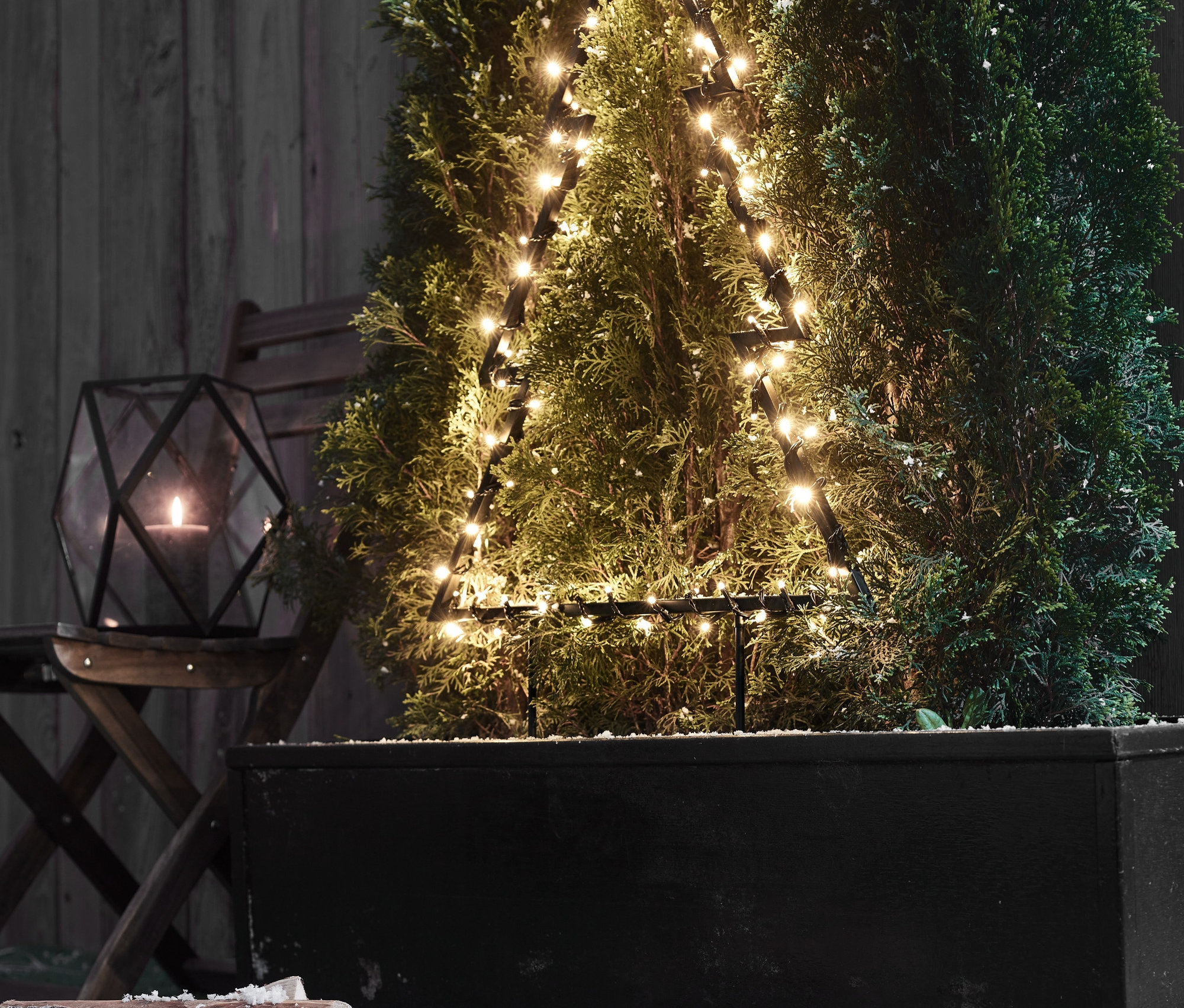 Siyah bir saksıda üçgen şeklinde büyük bir LED dış mekan çamı duruyor. Yanında bir fener ve odun var.