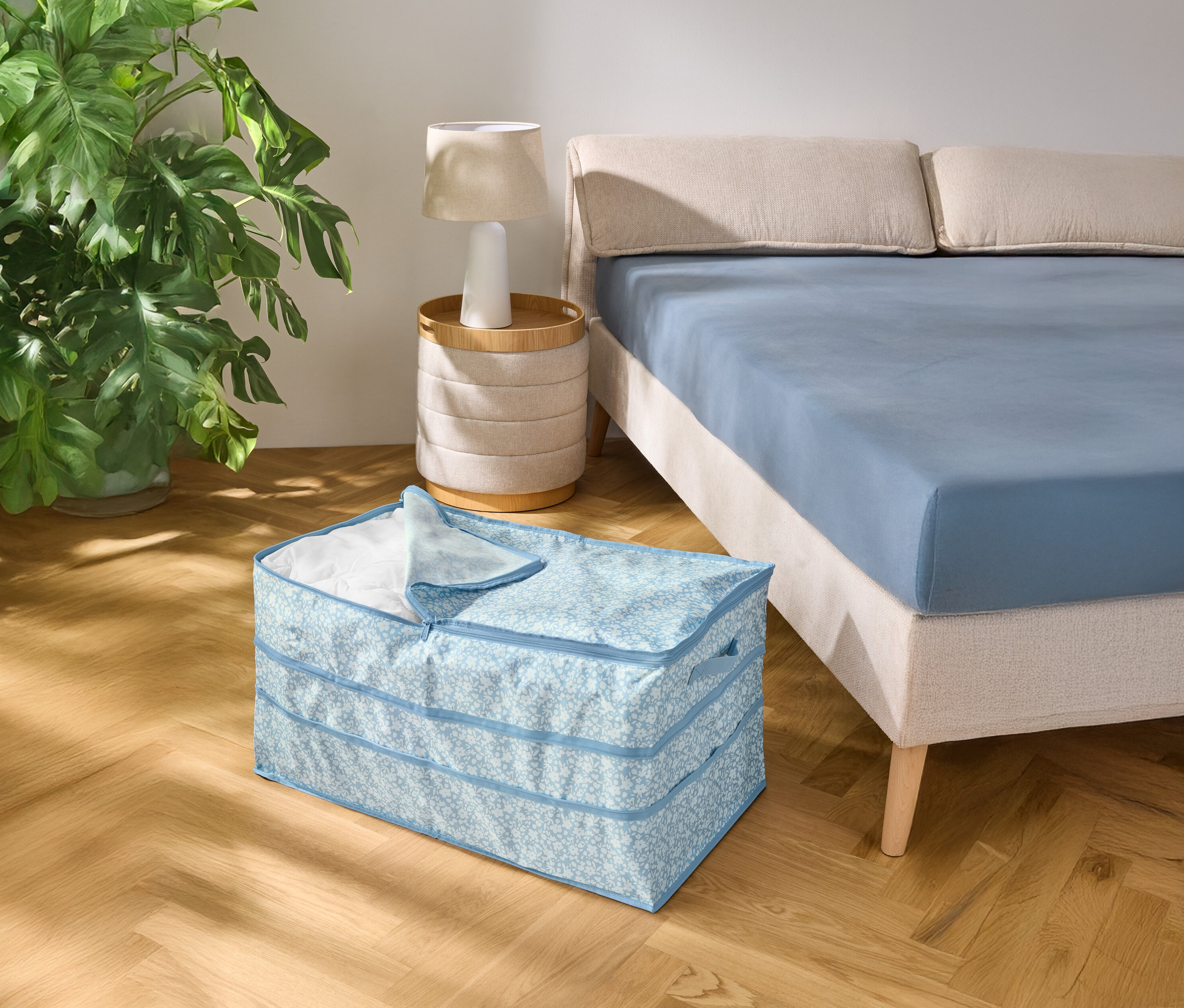 Ahşap zeminde, mavi çarşaflı bir yatağın yanında açık mavi desenli bir saklama kutusu duruyor.