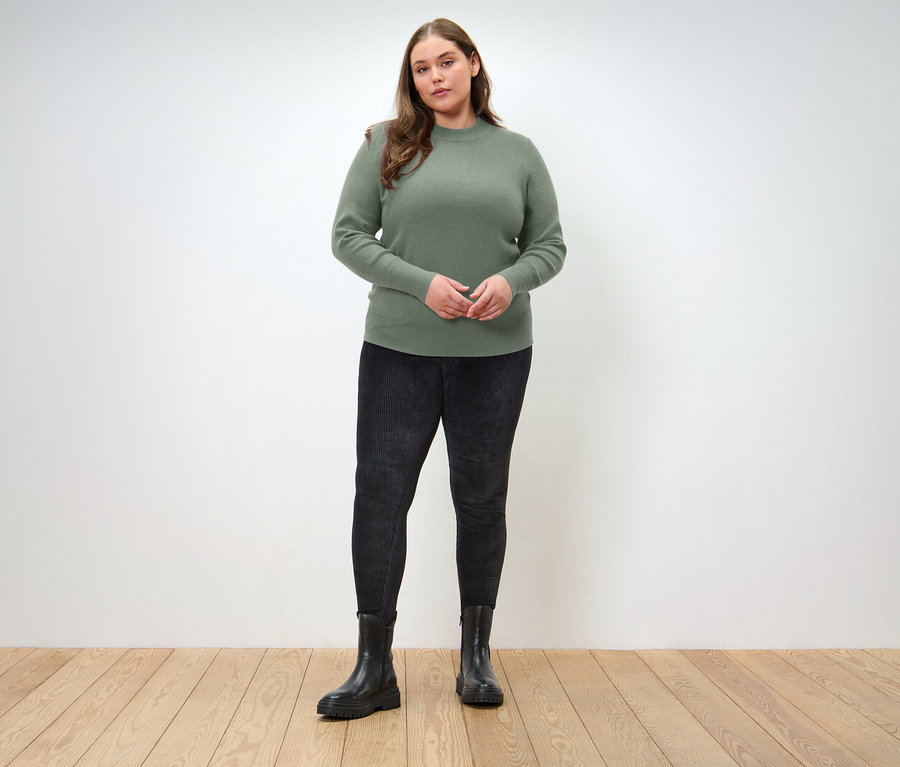 Ahşap zeminde duran bir kadın, üzerinde yeşil bir kazak, siyah kot pantolon ve siyah botlar var.