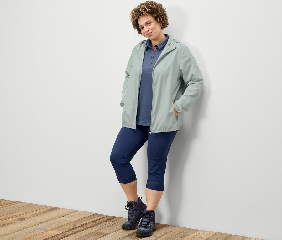 Duvara yaslanmış bir kadın, 3/4 trekking taytı, lacivert fonksiyonel polo tişört ve gri katlanabilir fonksiyonel mont giyiyor.