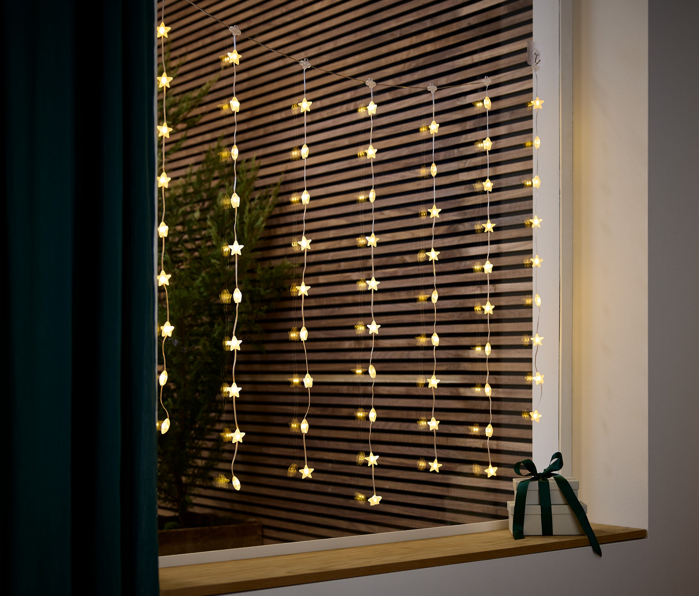 Yıldız şeklinde ışıklar, koyu yeşil kurdeleli bir hediyenin yanında pencerede asılı duruyor.