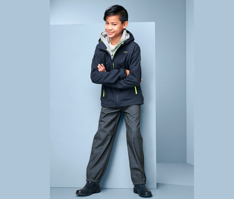 Bir çocuk kolları kavuşturmuş ayakta duruyor ve üzerinde kot görünümlü softshell pantolon, ceket ve kapüşonlu bir sweatshirt var.