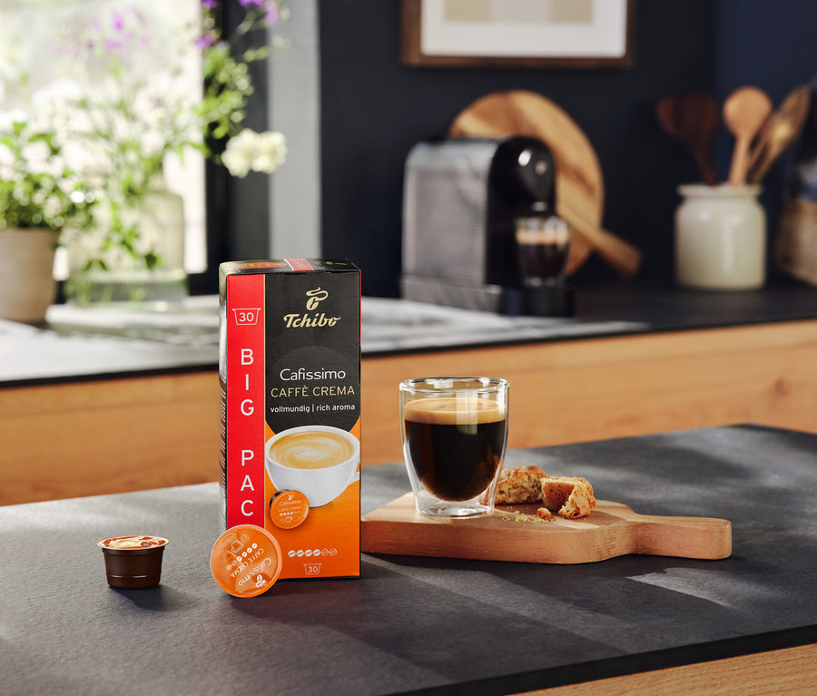 Bir paket Cafissimo Caffe Crema kapsülü, bir bardak kahve ve bir masa üzerinde kurabiyeler. Arka planda bir Cafissimo pure+ black kahve makinesi.