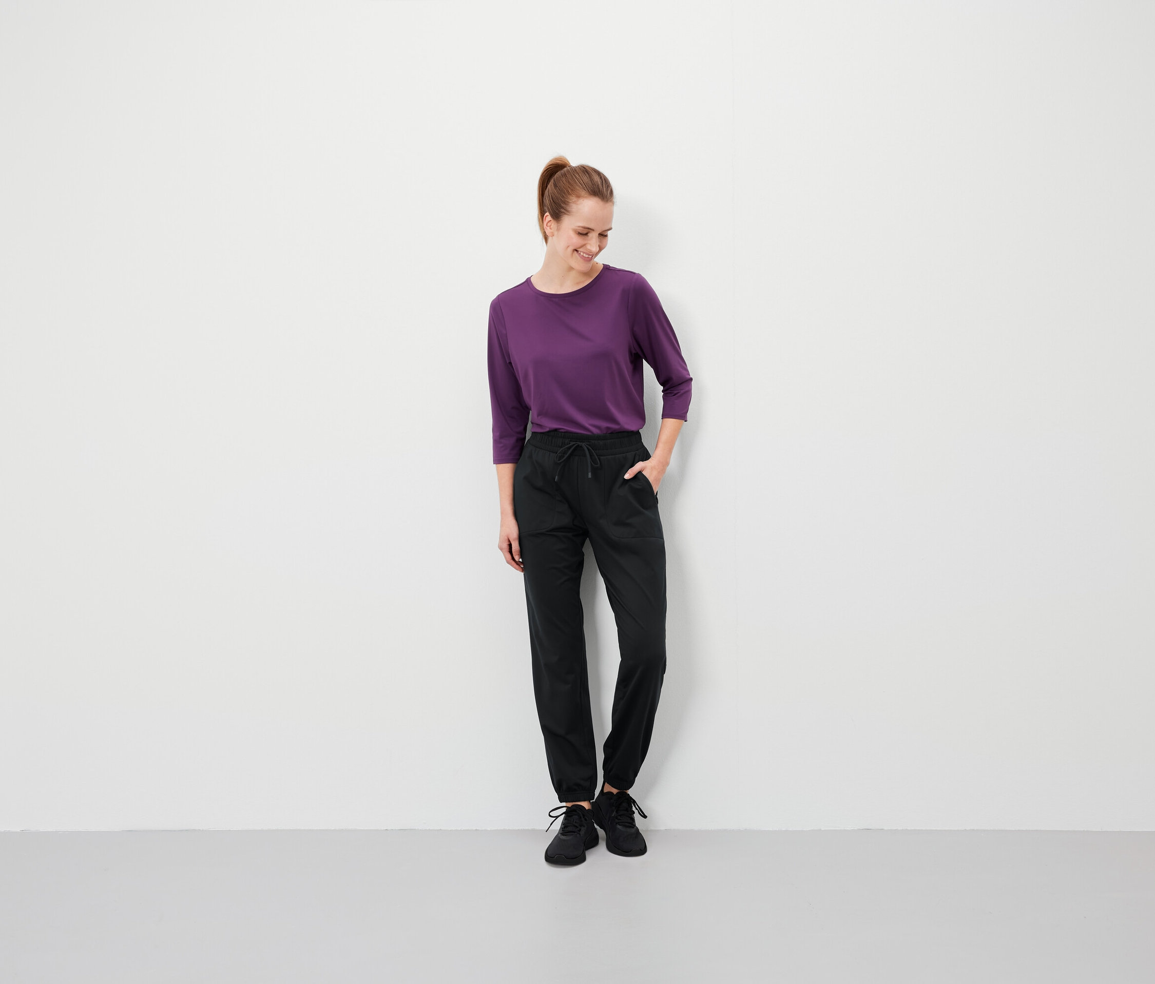 Kahverengi saçlı, at kuyruklu bir kadın, elleri siyah pantolonunun cebinde duruyor ve mor bir tişört giyiyor.