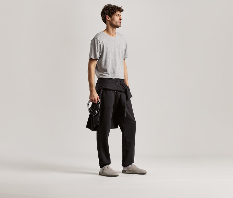 Adam, gri bir tişört ve siyah bir eşofman altı giyiyor. Elinde kulaklık tutuyor.