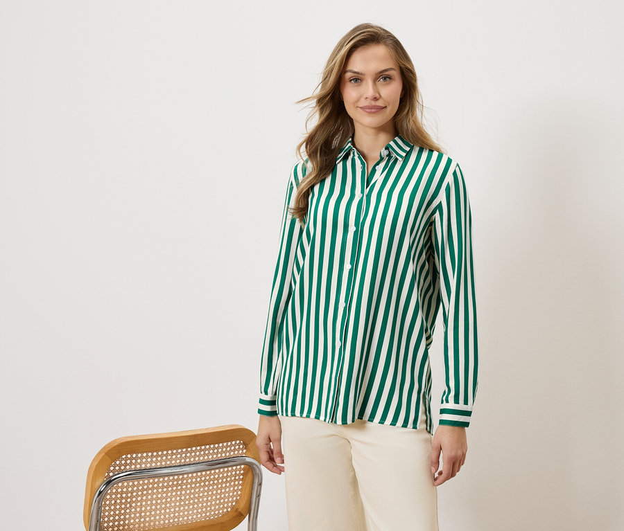 Kadın, yeşil ve beyaz çizgili bir gömlek ve bej pantolon giyiyor, yanında bir sandalye var.