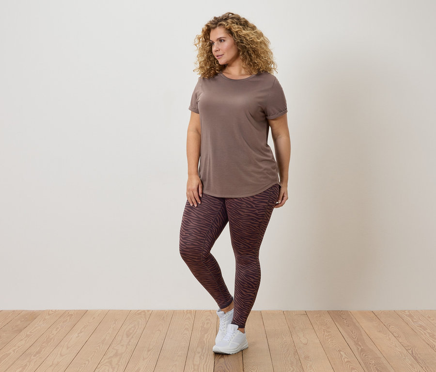 Kıvırcık saçlı, kahverengi tişörtlü ve desenli taytlı bir kadın duvarın önünde poz veriyor.