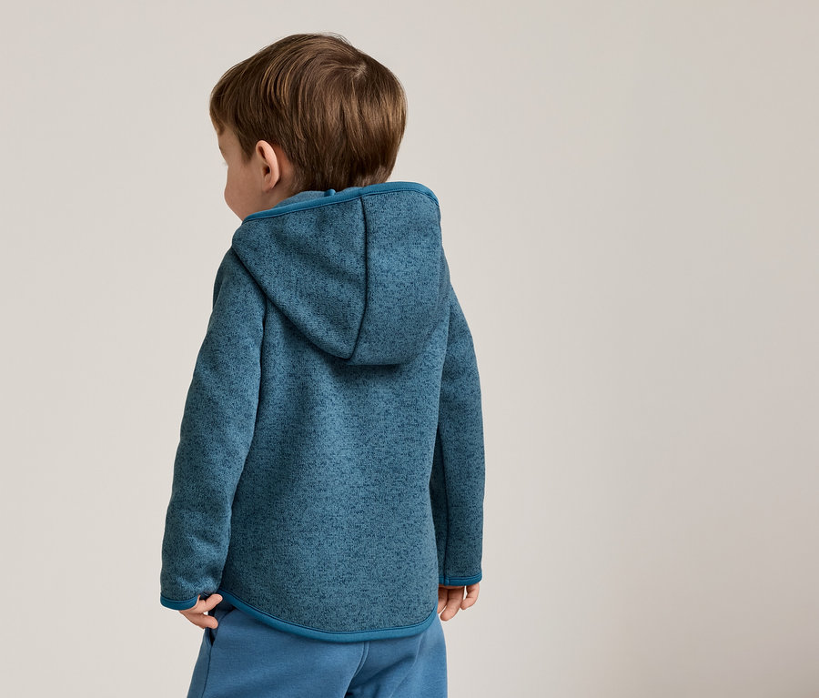 Kahverengi saçlı bir çocuğun arkadan görünüşü, mavi kırçıllı kapüşonlu bir sweatshirt ve mavi pantolon giyiyor.
