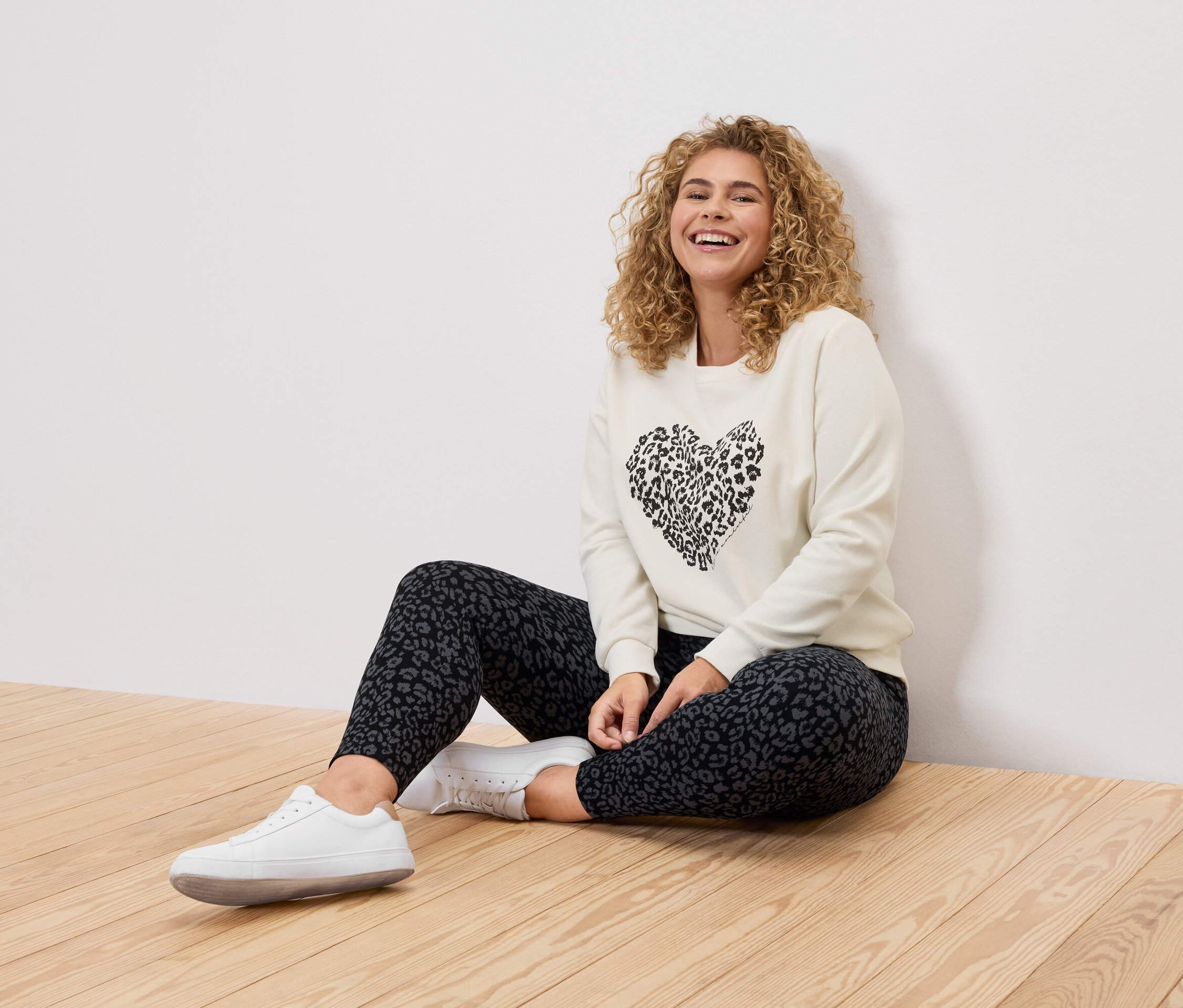 Kıvırcık saçlı bir kadın, krem rengi bir kazak, leopar desenli tayt ve beyaz spor ayakkabı giyerek ahşap bir zeminde oturuyor.