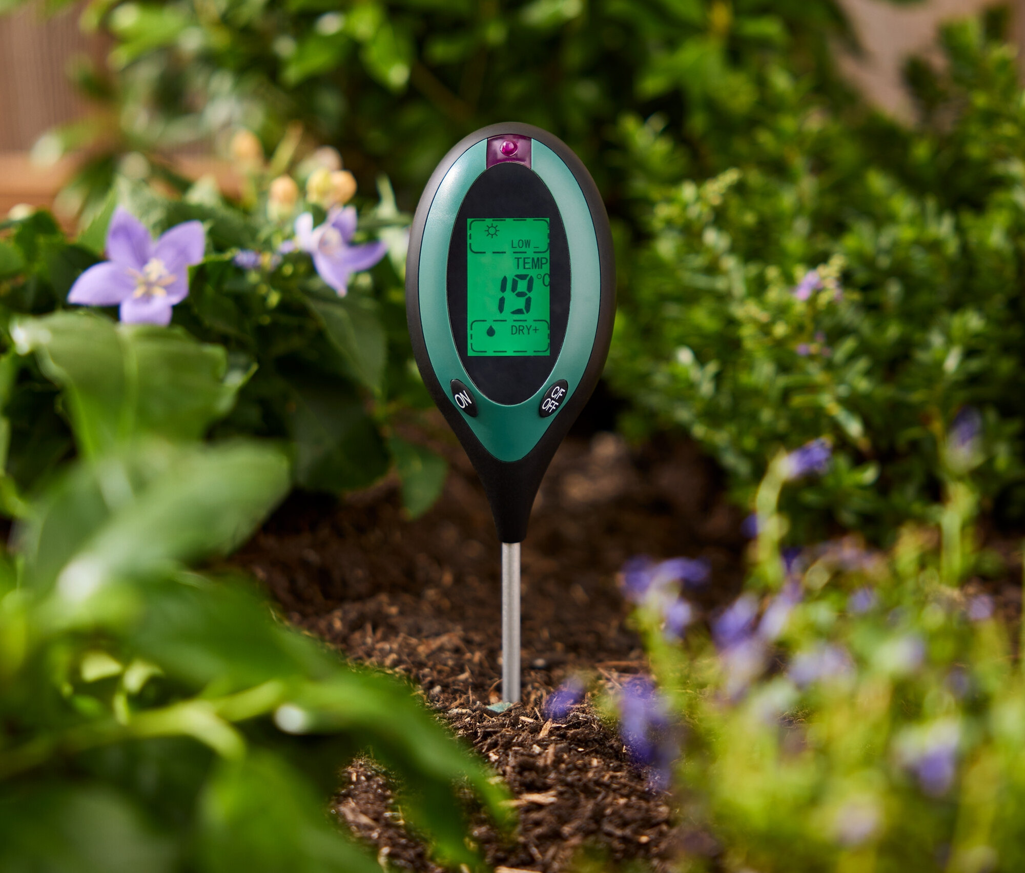 Yeşil bir toprak test cihazı, bitkilerin arasındaki toprağa saplanmış.