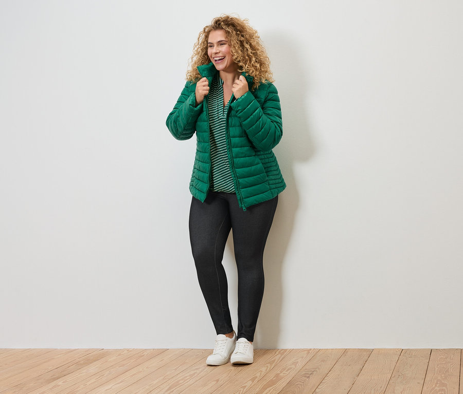 Kıvırcık saçlı, yeşil şişme montlu, çizgili bluzlu, koyu renk taytlı ve beyaz spor ayakkabılı kadın.