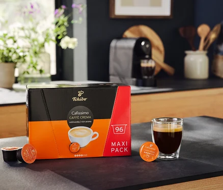 Bir paket Cafissimo Caffè Crema vollmundig 96 kapsül, bir bardak kahvenin yanında duruyor. Arka planda bir Cafissimo pure+, siyah kahve makinesi var.