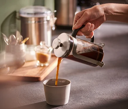 Bir el, 300 ml gümüş French Press'ten bir fincana kahve döküyor.