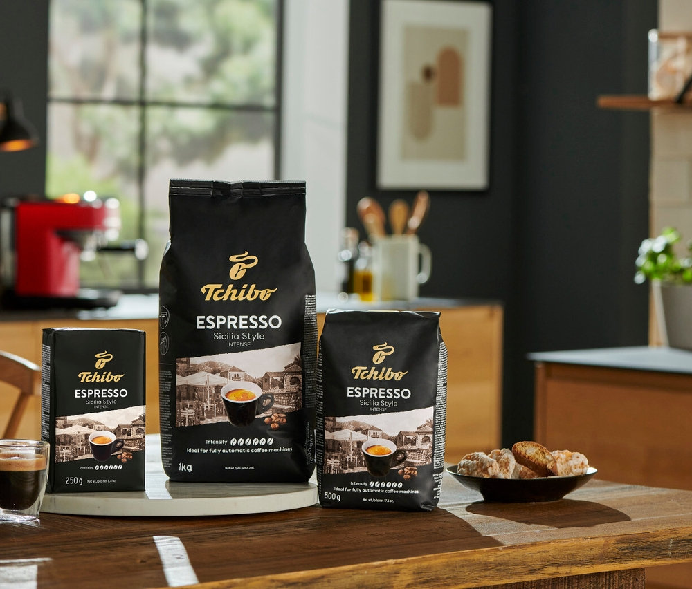 Tahta bir masanın üzerinde bir fincan espressonun ve bir tabak kurabiyenin yanında üç paket Tchibo Espresso Sicilia Style kahvesi (1 kg, 500 g, 250 g) duruyor.