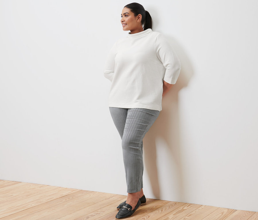 Duvara yaslanmış bir kadın krem rengi 3/4 kollu sweatshirt, streç pantolon ve deri loafer ayakkabı giyiyor.