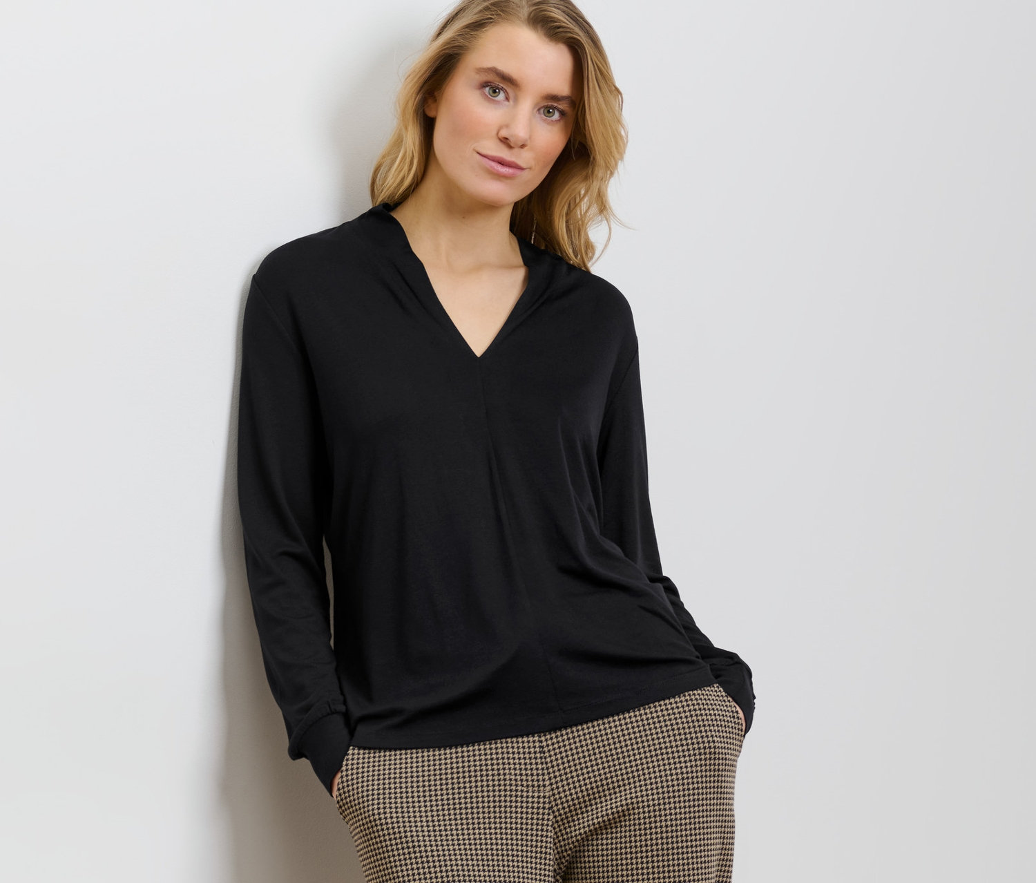 Duvara yaslanmış bir kadın, siyah V yakalı bir bluz ve kareli pantolon giyiyor.