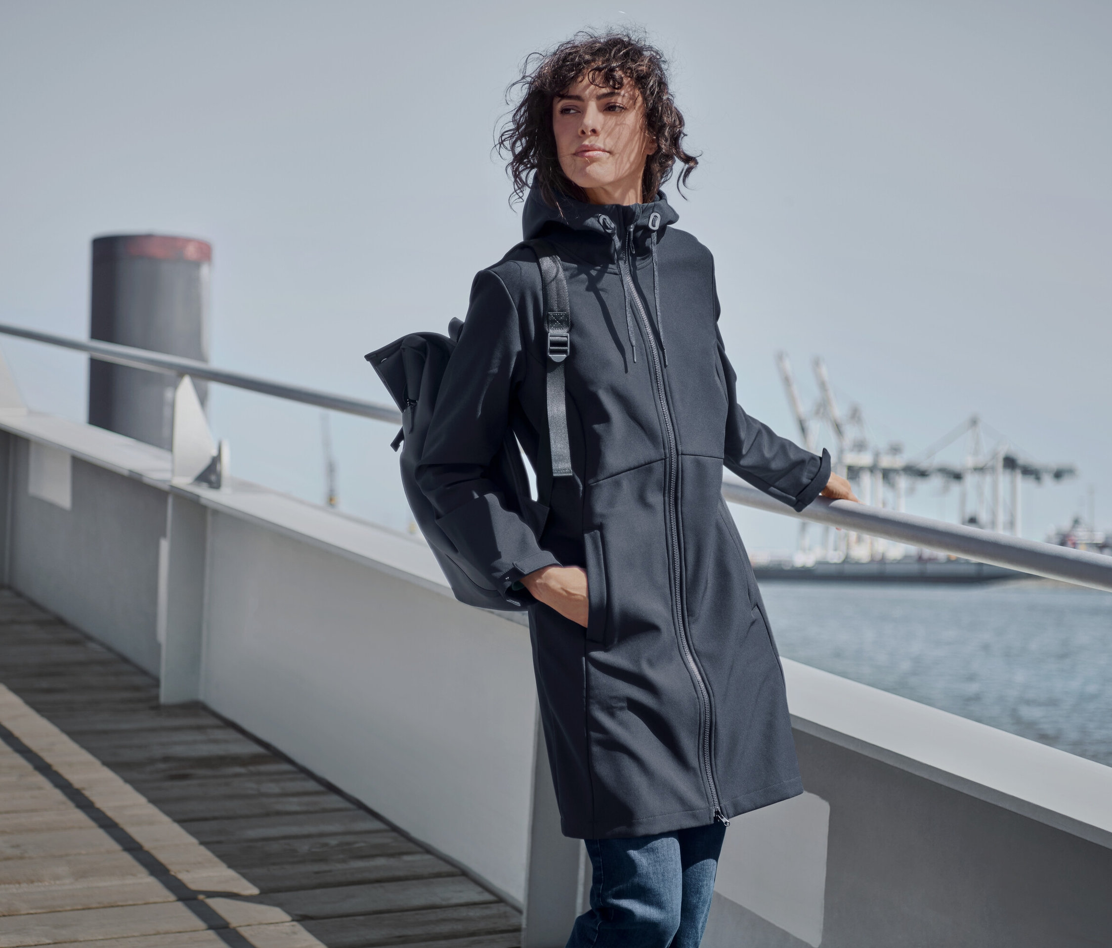 Korkuluğa yaslanan kadın lacivert softshell palto, düz kesim kot pantolon ve su geçirmez sırt çantası giyiyor.