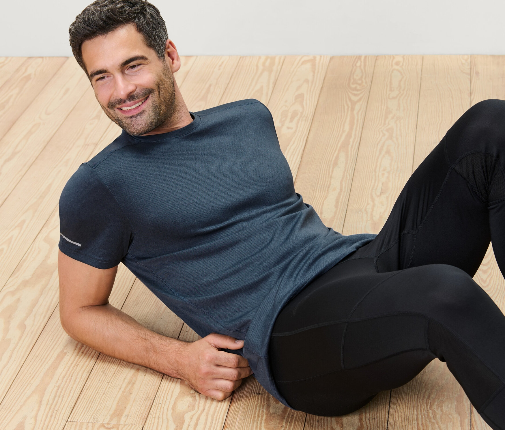 Adam ahşap zeminde uzanmış, kırçıllı mavi fonksiyonel tişört giyiyor.