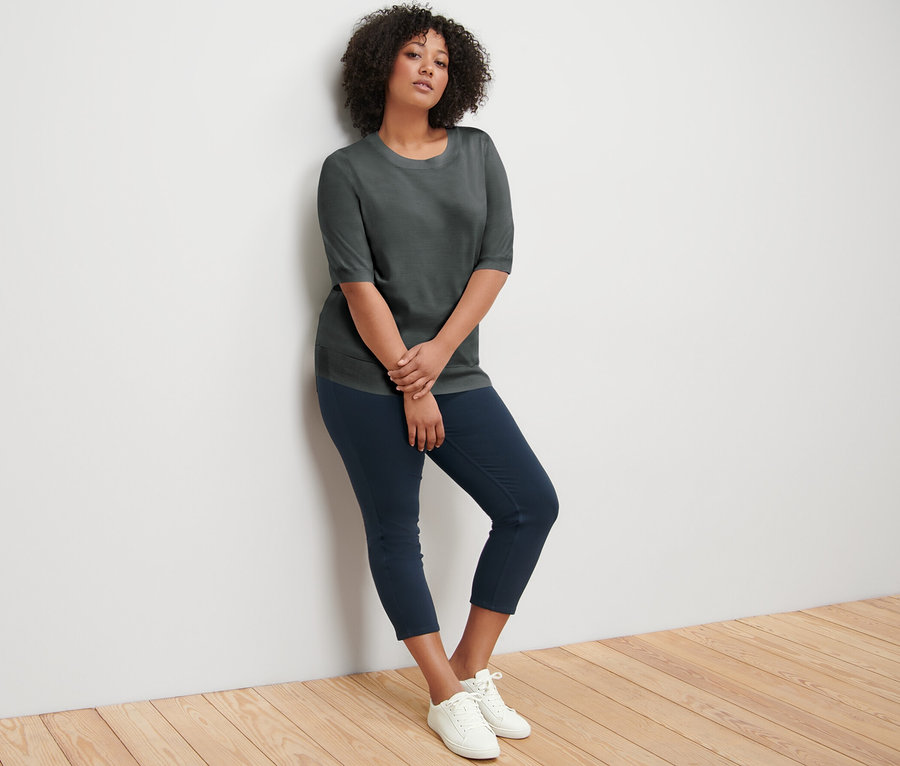 Duvara yaslanmış bir kadın, yeşil ince triko kazak, lacivert pantolon ve beyaz spor ayakkabı giyiyor.