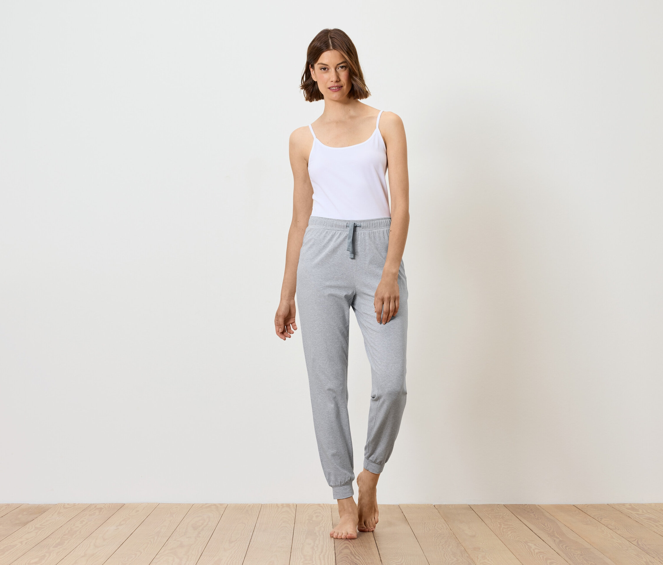 Kadın beyaz atlet ve gri eşofmanla ayakta duruyor, çıplak ayakla.