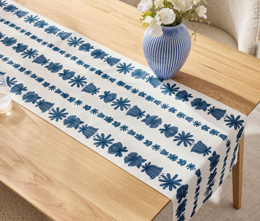 Bej renkli ahşap masa üzerinde mavi-beyaz bir masa örtüsü, beyaz çiçekli mavi bir vazo ve bir bardak.