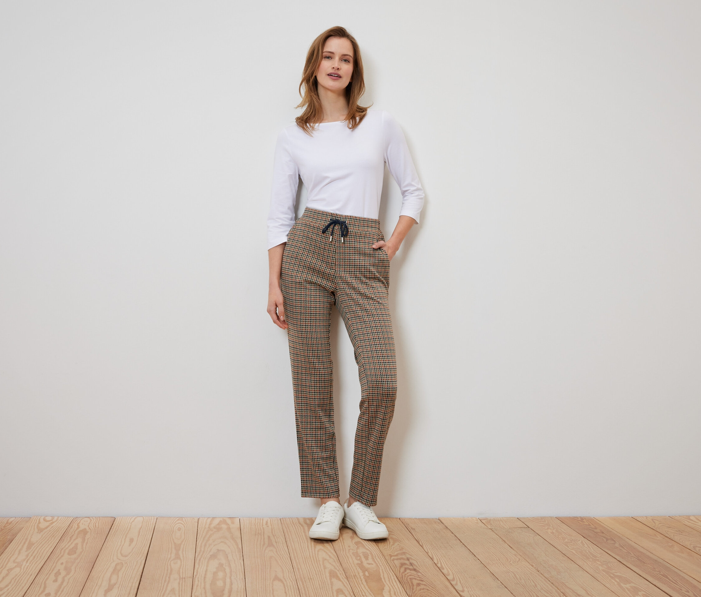 Kareli pantolon ve beyaz gömlek giyen bir kadın ayakta duruyor ve poz veriyor.