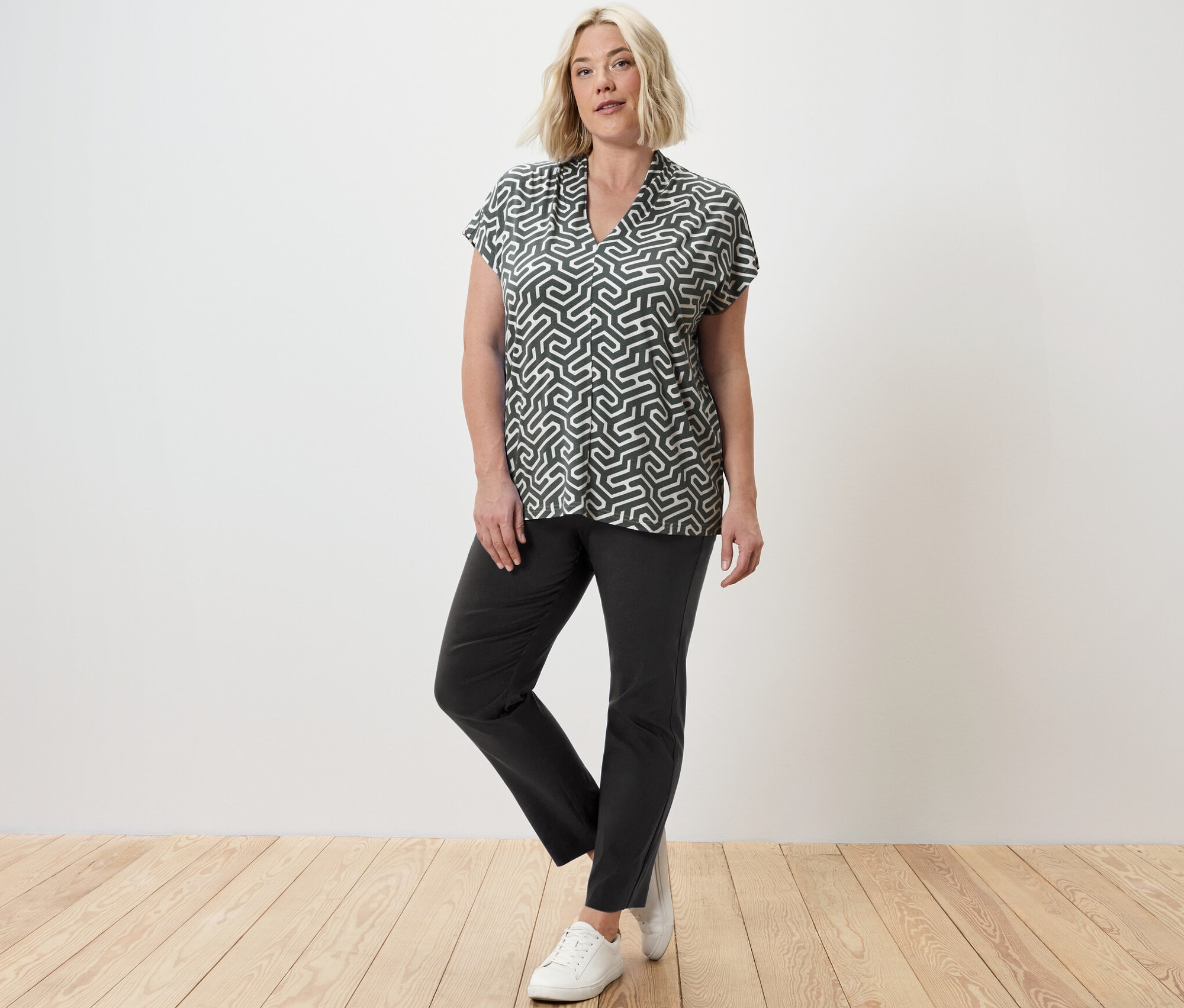 Sarışın bir kadın, yeşil V yakalı bluz ve streç pantolonla poz veriyor.