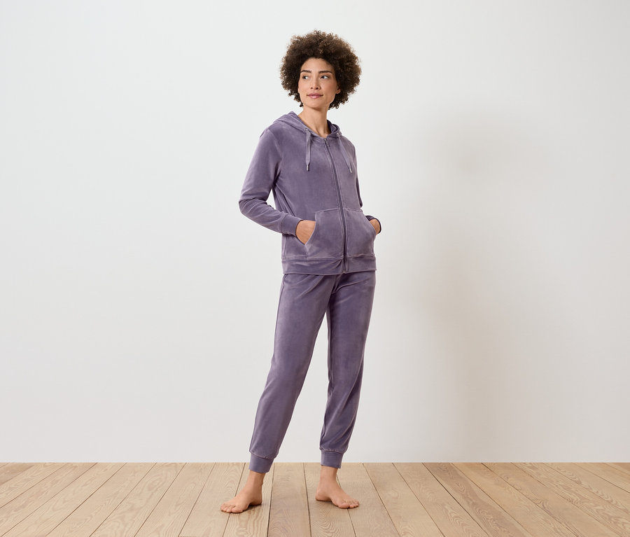 Kadın ayakta duruyor, üzerinde mor kapüşonlu ve eşofman altı var.