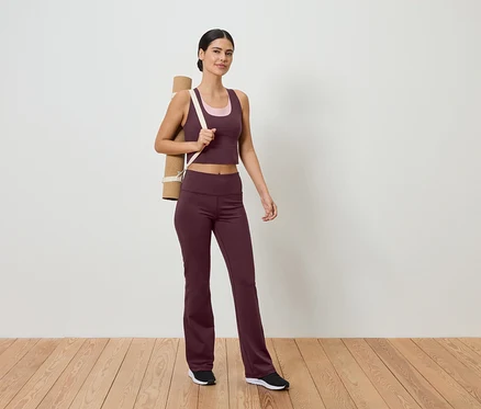 Spor kıyafetli, omzunda yoga matı olan bir kadın, beyaz duvar önünde ahşap zeminde duruyor.