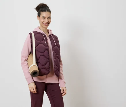 Kadın, pembe kapüşonlu sweatshirt ve bordo tayt üzerine bordo kapitone yelek giyiyor, omzunda bir yoga matı var.