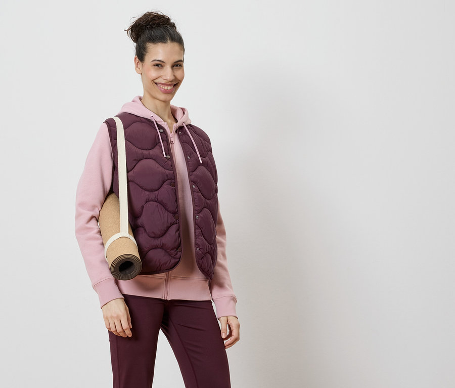 Kadın, pembe kapüşonlu sweatshirt ve bordo tayt üzerine bordo kapitone yelek giyiyor, omzunda bir yoga matı var.