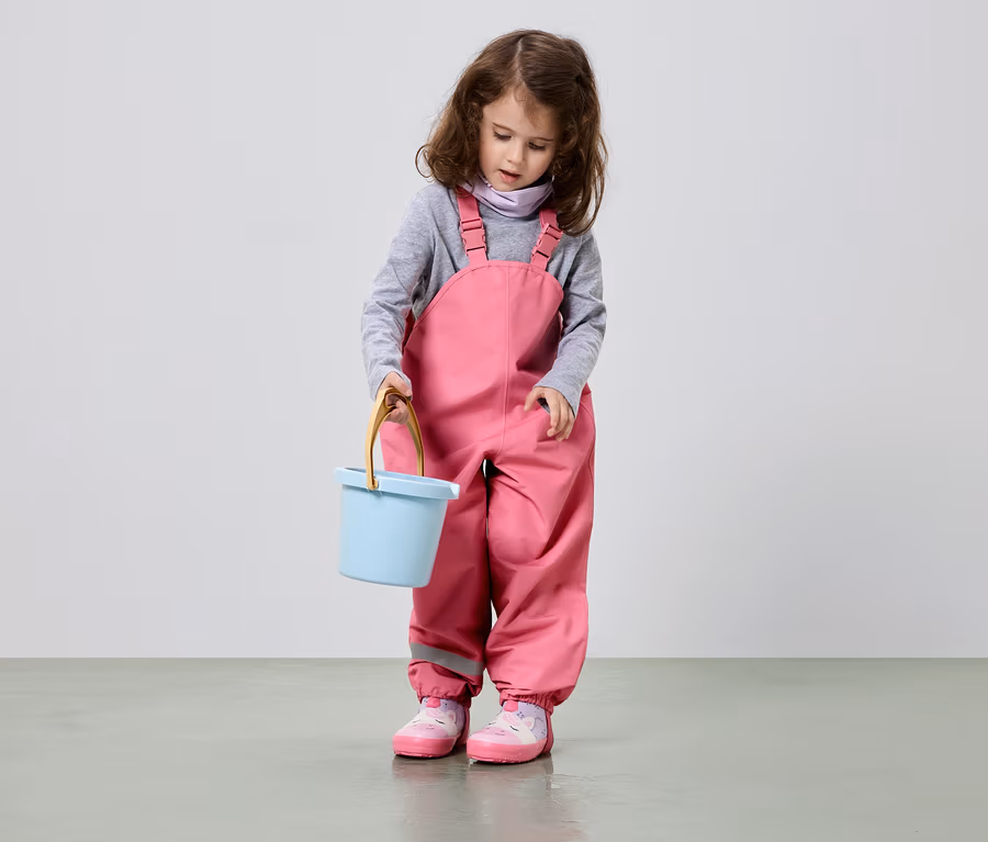 Kahverengi bukleli saçları olan bir kız çocuğu açık mavi bir kova tutuyor, pembe bir salopet ve pembe ayakkabılar giyiyor.