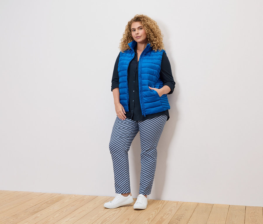 Kıvırcık saçlı, mavi yelekli, koyu renk gömlekli ve desenli pantolonlu bir kadın beyaz bir duvarın önünde duruyor.