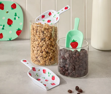 Fotoğrafta granola ve kuru üzüm dolu iki cam kap ve aralarında bir süt şişesi yer alıyor. Kapların üzerinde çilek desenli kürekler bulunuyor.