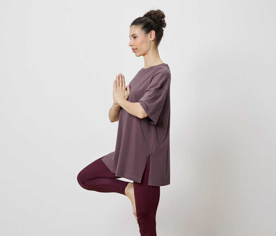 Kahverengi üst ve taytlı bir kadın, tek bacak üzerinde yoga pozu yapıyor.