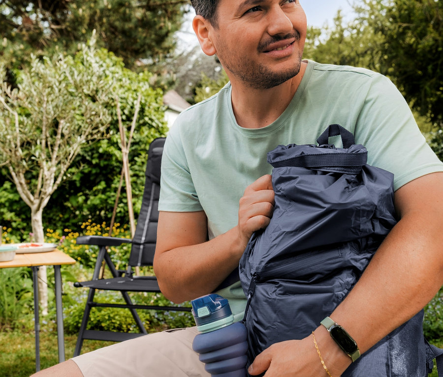 Nane yeşili Outdoor Tişört giyen bir adam bahçede oturuyor, koyu mavi Su Geçirmez Sırt Çantası ve mavi katlanabilir bir su şişesi tutuyor.