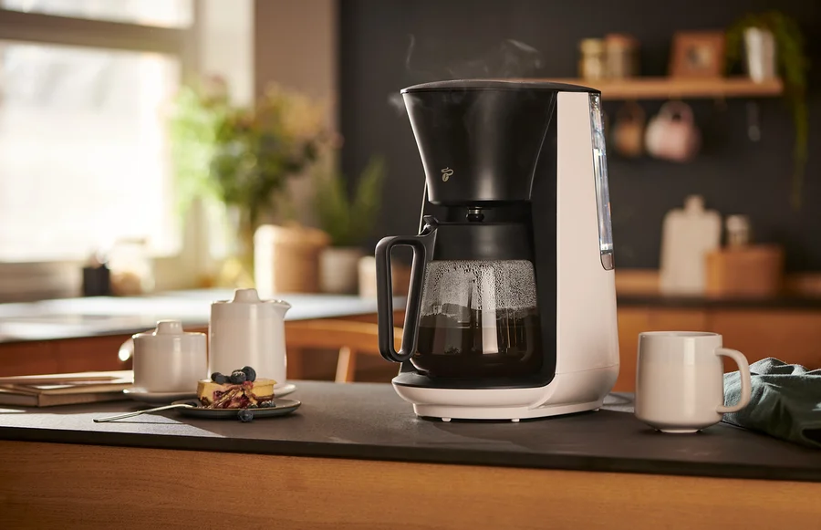 Beyaz »Let's Brew« Filtre Kahve Makinesi bir fincan, sütlük, şekerlik ve bir tabak kekin yanında bir masanın üzerinde duruyor.