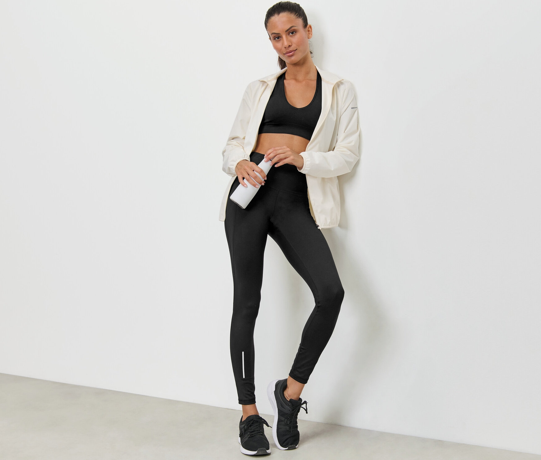 Duvara yaslanmış bir kadın elinde su şişesi tutuyor, sporcu sütyeni, ceket ve su itici spor tayt giyiyor.