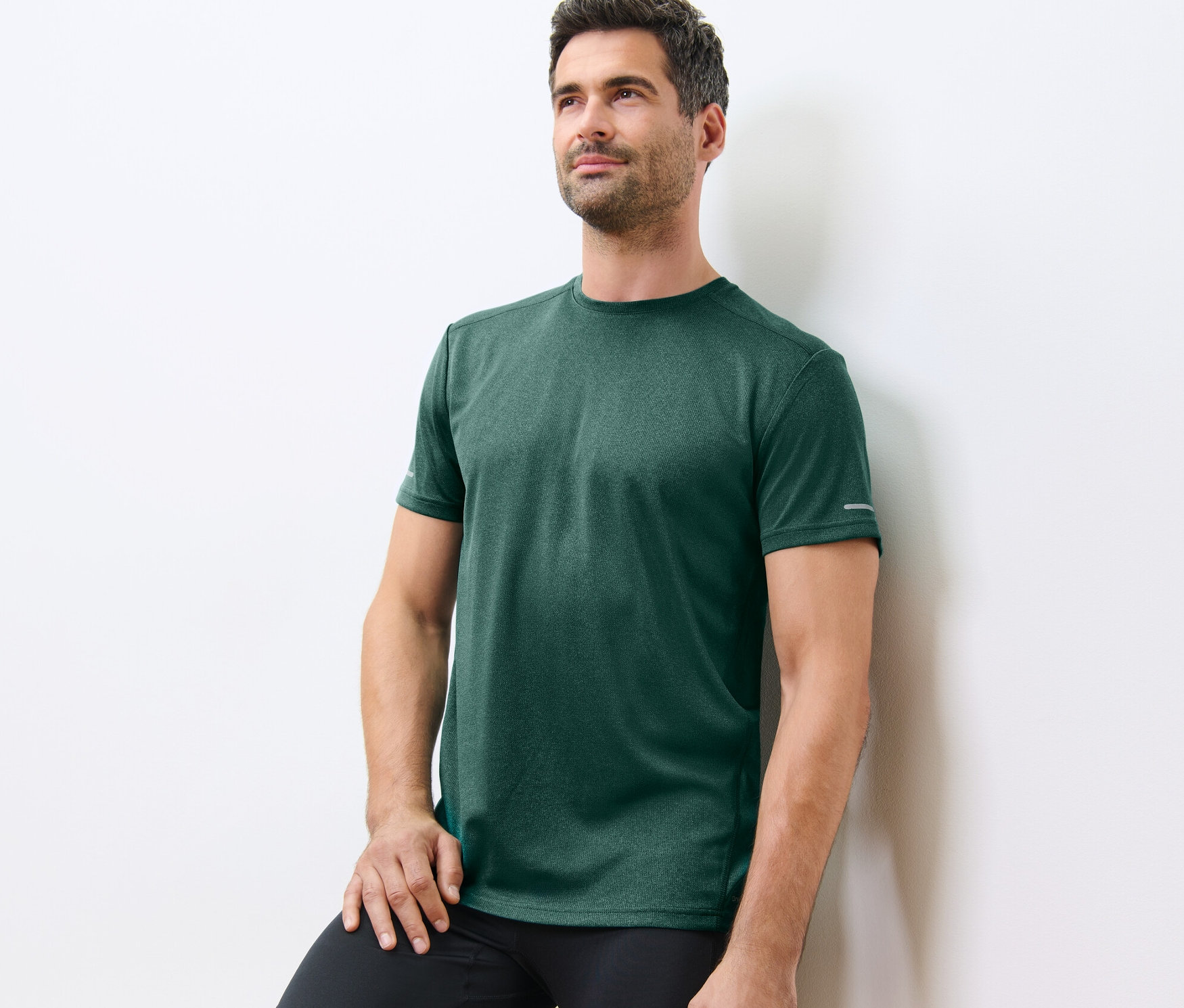 Beyaz bir duvara yaslanan adam kırçıllı yeşil bir fonksiyonel tişört giyiyor.