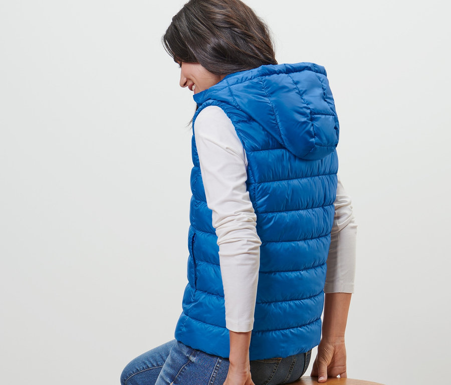 Kadın ahşap taburede oturuyor ve mavi kapüşonlu kapitone yelek giyiyor.