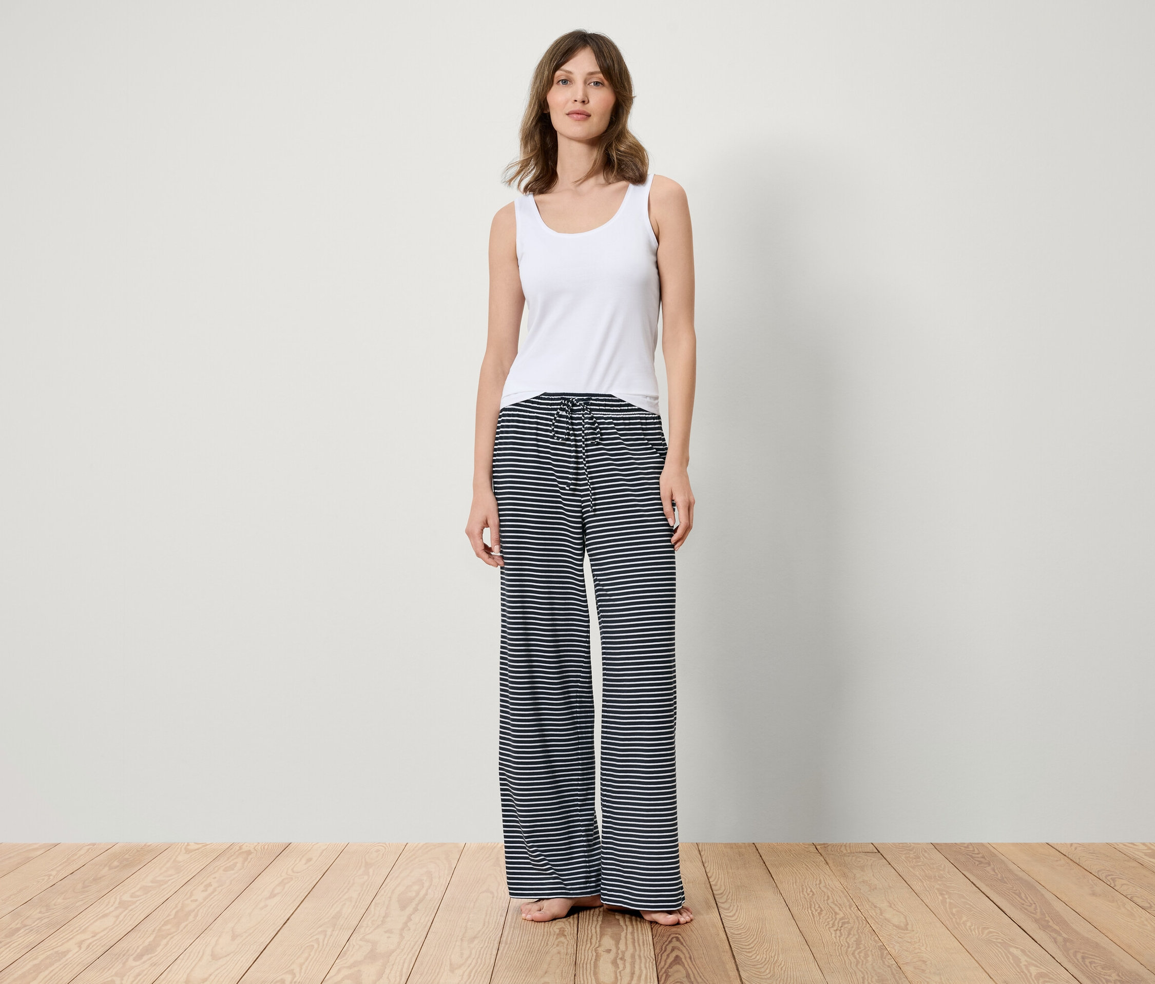 Ahşap zeminde duran bir kadın, beyaz bir atlet ve mavi beyaz çizgili bir pantolon giyiyor.
