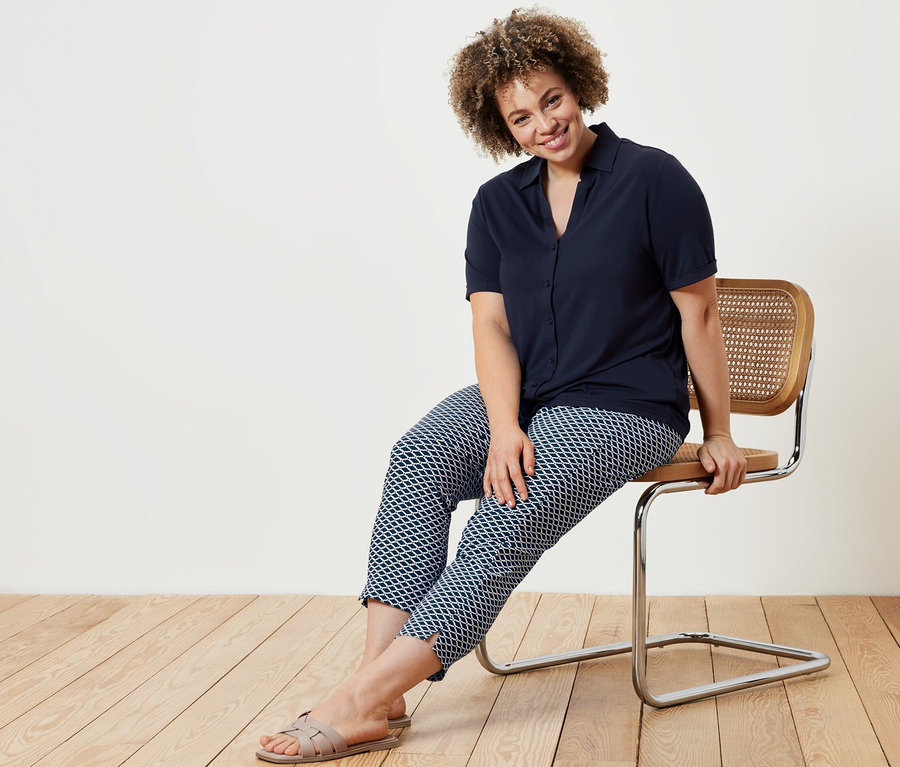 Bir kadın sandalyede oturuyor, üzerinde lacivert düğmeli bluz ve baskılı 7/8 streç pantolon var.
