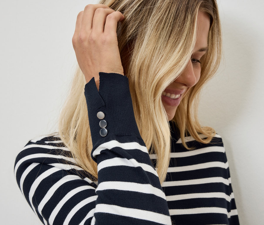 Sarışın bir kadın, kolunda düğmeleri olan çizgili bir kazak giyerken eliyle saçını düzeltiyor.