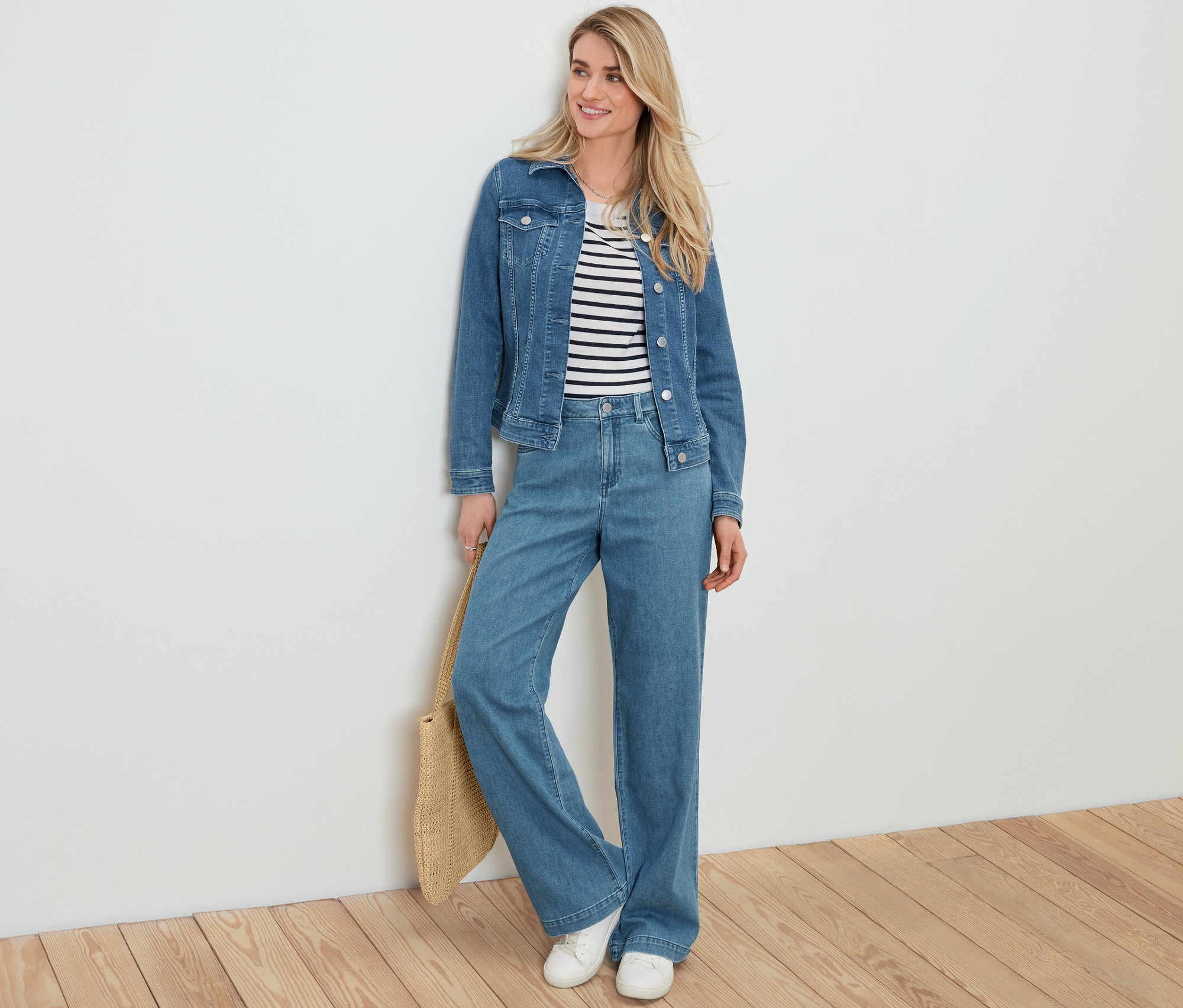 Sarışın bir kadın beyaz bir duvara yaslanmış, üzerinde kot ceket, çizgili bir kazak, palazzo kot pantolon ve elinde hasır görünümlü bir alışveriş çantası var.