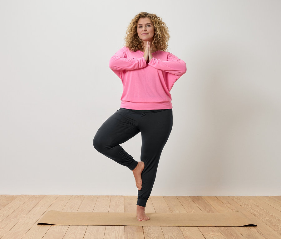 Kıvırcık saçlı kadın bej bir mat üzerinde yoga yapıyor, pembe bir üst ve gri pantolon giyiyor.